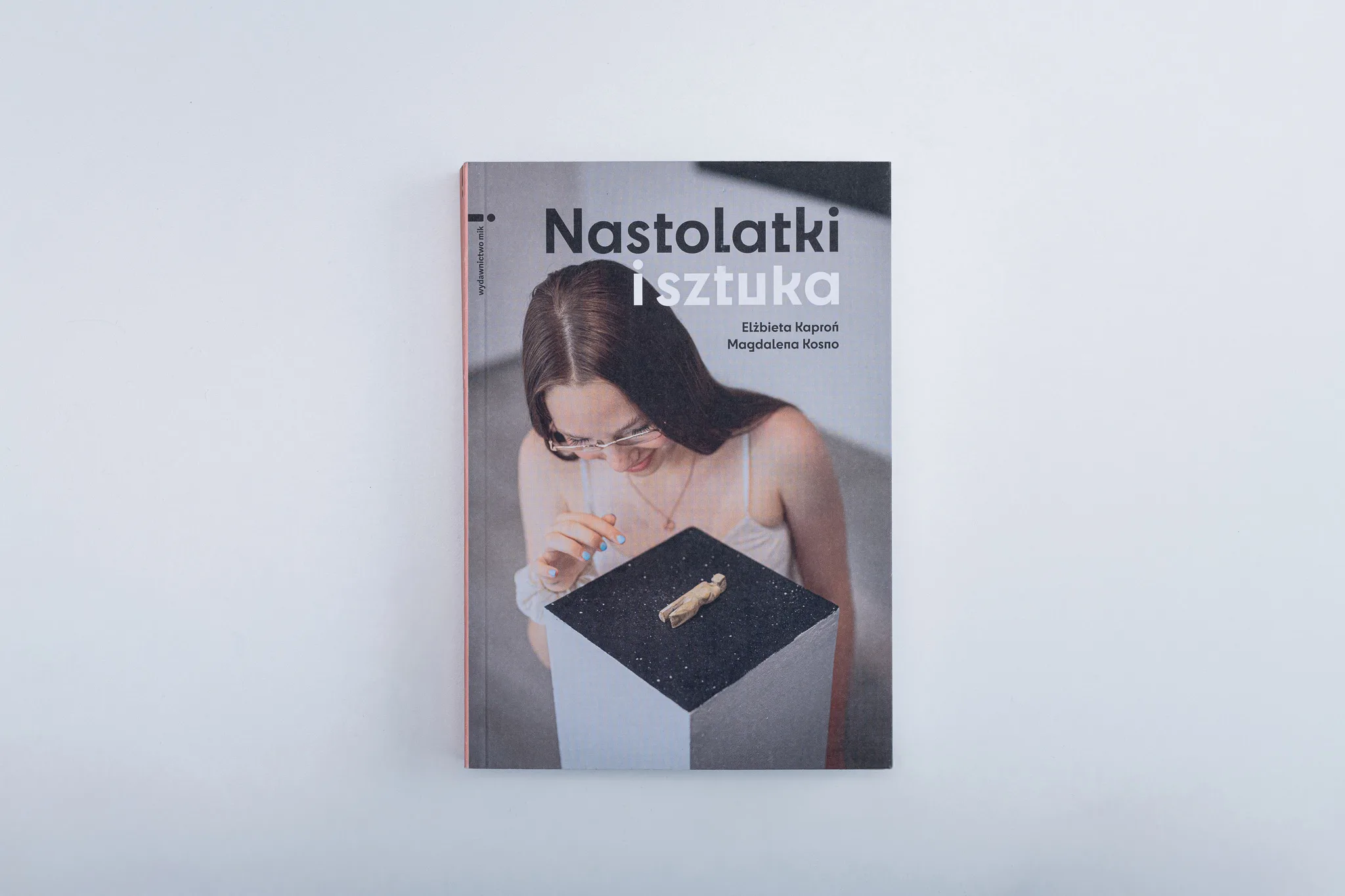 Okładka książki z fotografią młodej, długowłosej dziewczyny przyglądającej się ciekawemu, małemu eksponatowi na postumencie