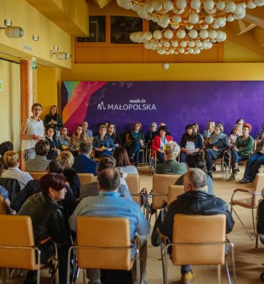 sala wypełniona ludźmi siedzącymi na krzesłach - przy ścianie widać fioletową, dużą grafikę z logotypem Małopolska