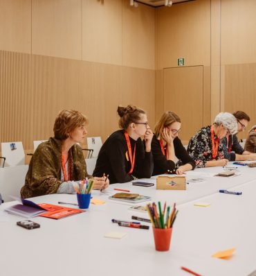 długi stół, przy którym siedzą ludzie i dyskutują, notują