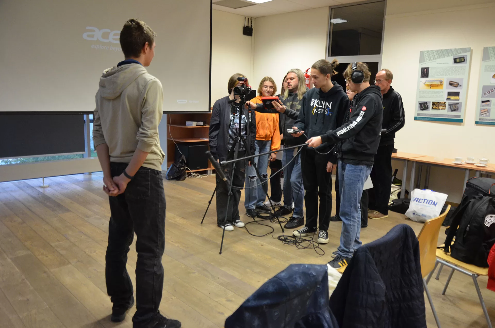 Grupa młodzieży realizuje nagranie aparatem na statywie i mikrofonem - przed obiektywem występuje chłopak