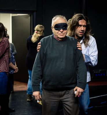 zdjęcie mężczyzny z zasłoniętymi oczami, prowadzonego przez drugiego mężczyznę